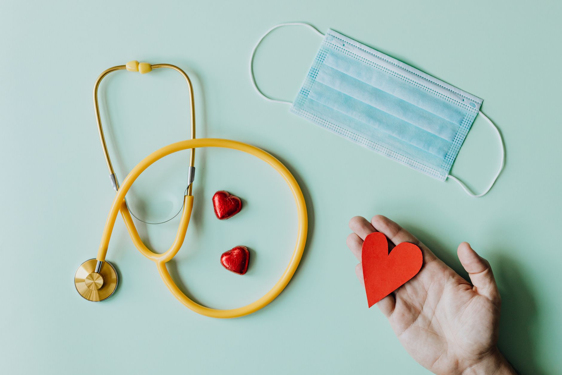 Top view of crop anonymous person hand with red paper heart on table with stethoscope and medical mask for coronavirus preven