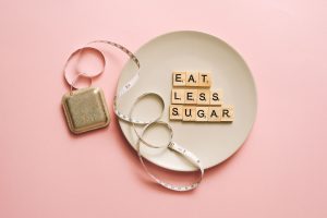 Flat lay of Scrabble tiles saying 'Eat Less Sugar' on a plate with a tape measure on a pink background. — Detox Diets - What