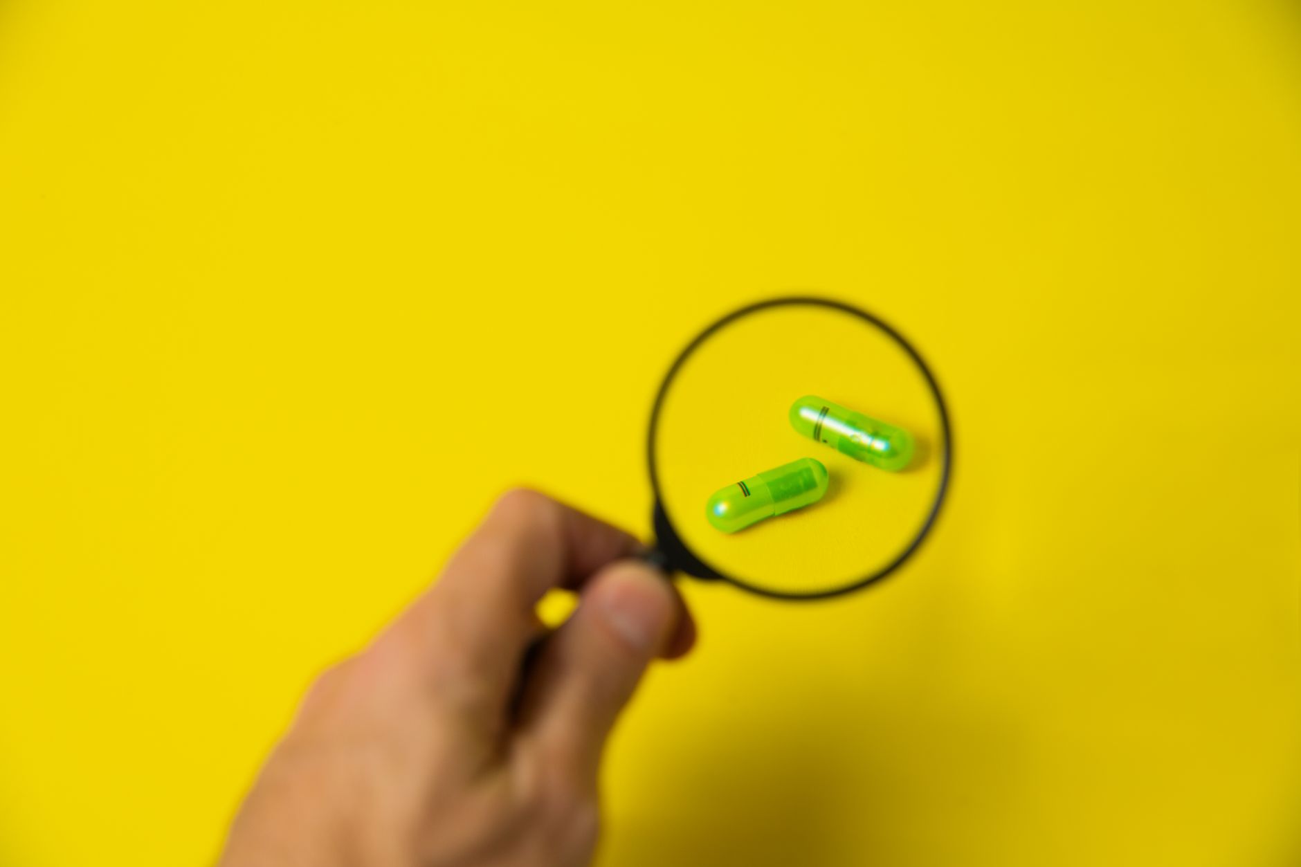From above of crop unrecognizable male holding magnifying glass over green pills on yellow background in studio — Get to Know