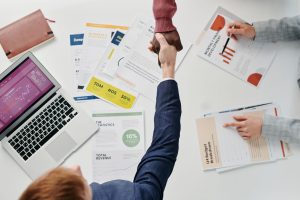 Business professionals engaging in a handshake over a desk filled with financial documents and a laptop. — Simplify Complianc