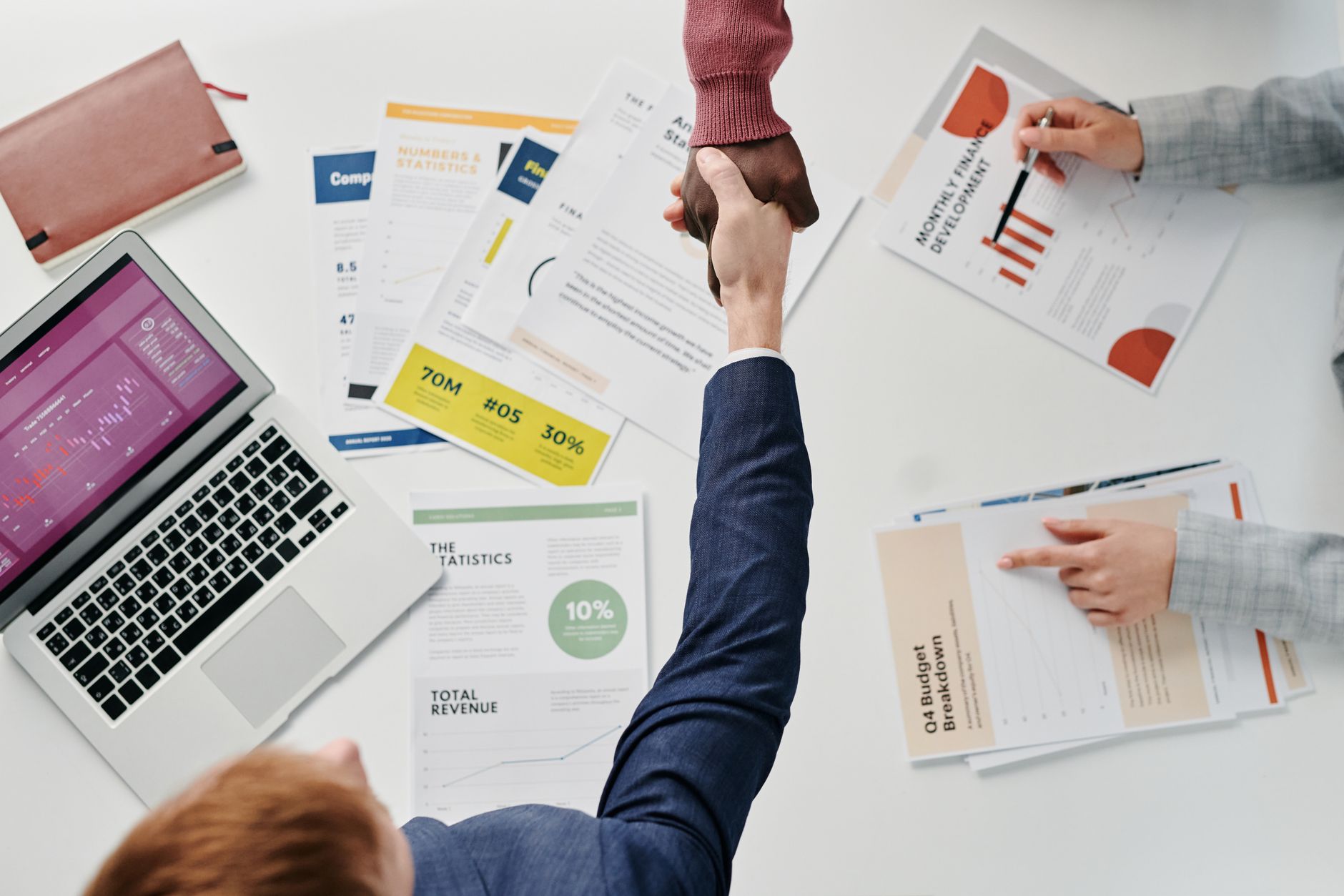 Business professionals engaging in a handshake over a desk filled with financial documents and a laptop. — Simplify Complianc