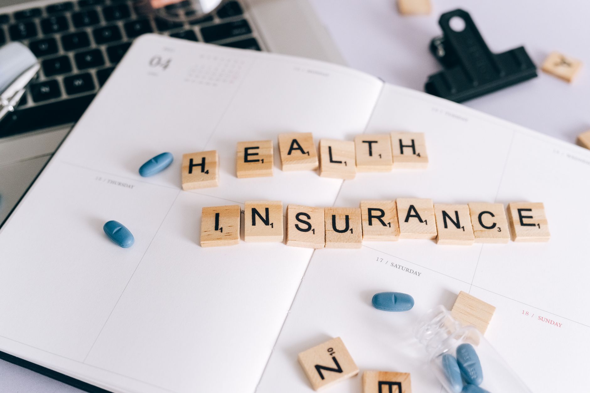 Scrabble tiles spelling 'Health Insurance' on planner with pills and laptop, symbolizing healthcare planning. — Tips to help