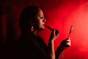 Elegant woman applying lipstick in dramatic lighting against a deep red background. — Where Do Acne Scars Occur and How to Cu