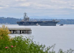A large aircraft carrier sailing in a calm body of water with a wooden pier in the foreground.